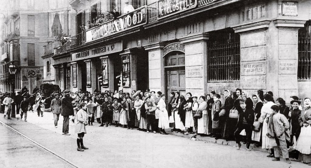 Fotografía antigua, decenas de personas hacen cola frente a un edificio.