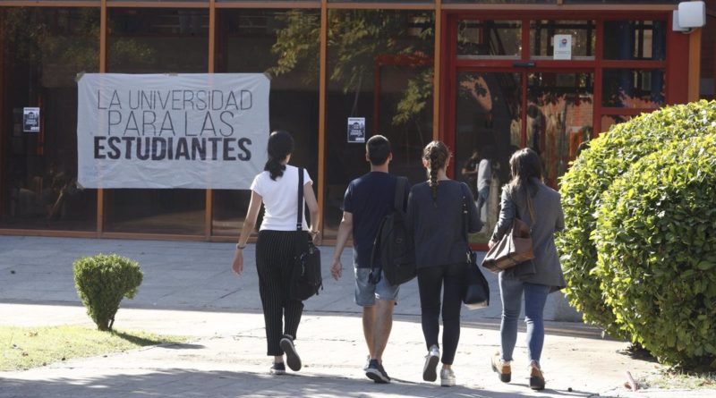Cuatro estudiantes de espaldas se dirigen a la entrada de una universidad junto a la cual hay una pancarta en la que se puede leer "La universidad para las estudiantes"